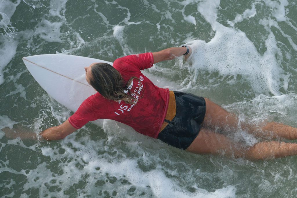 Karina Petroni paddles out for her heat in the WaveMasters pro division. She wears a red rash guard and the surf is foamy. The image is shot from overhead.