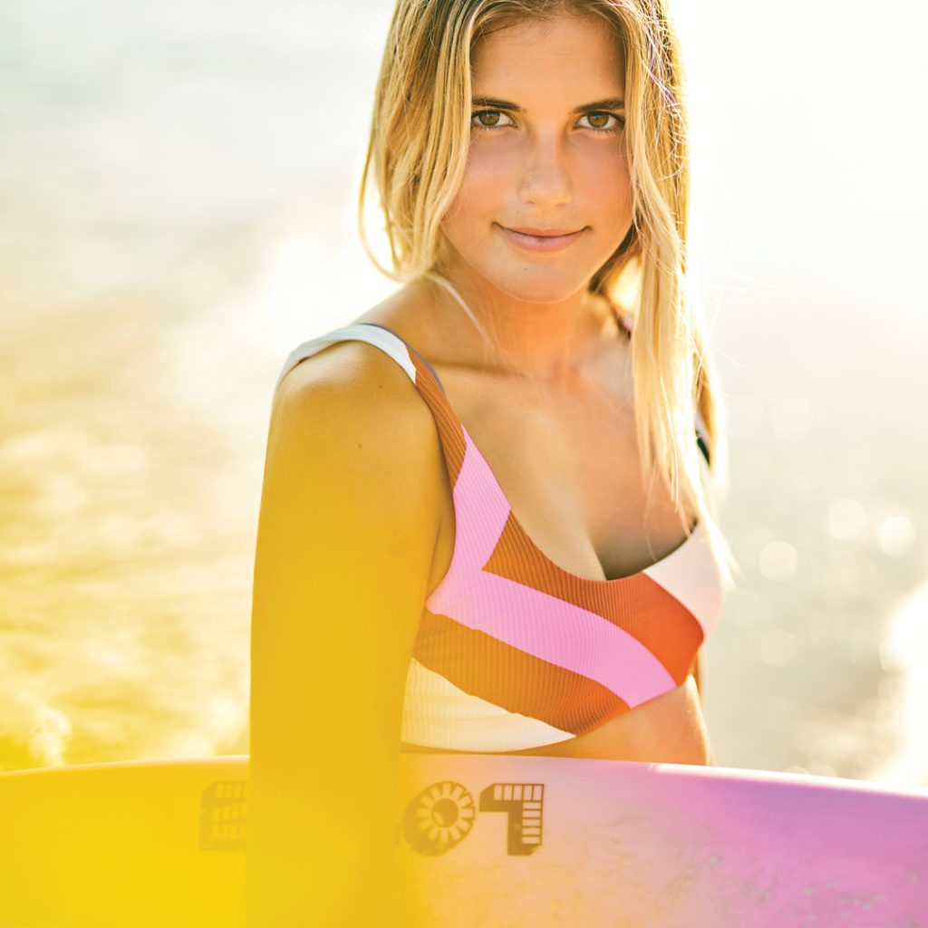 Benedetto is illuminated by the sunshine. She looks at the camera in the close-up shot while holding her surfboard. 