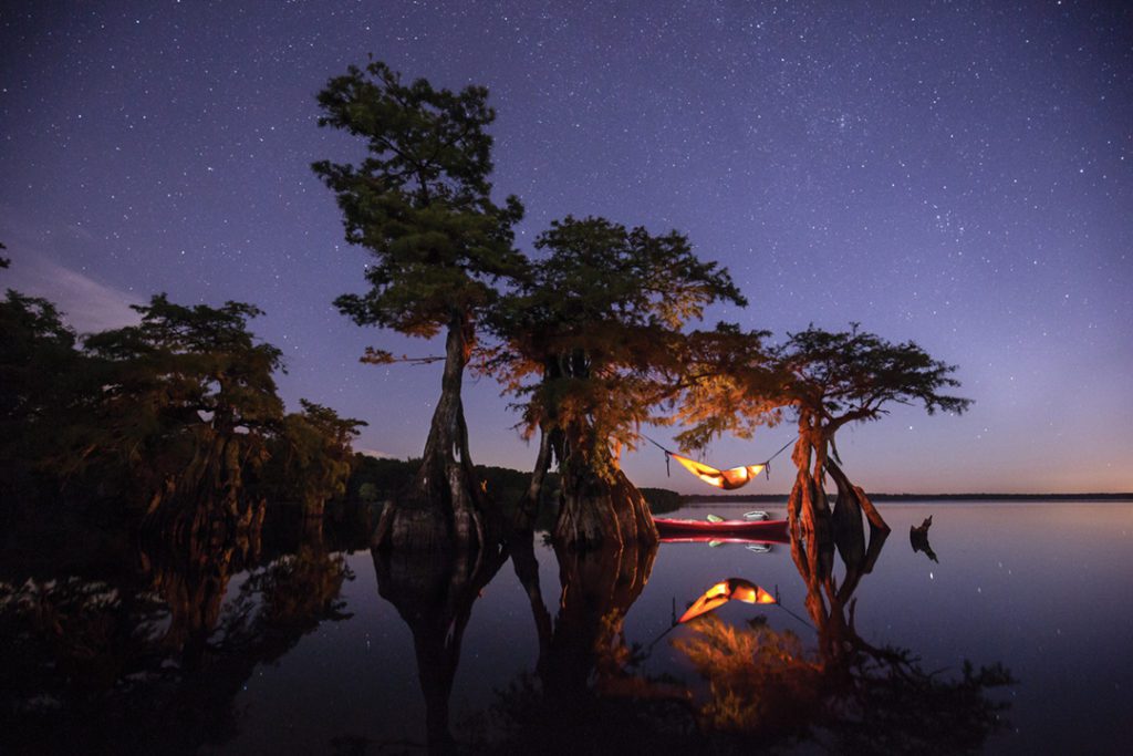 Image of a man in a lighted hammock underneath a tree on a lake