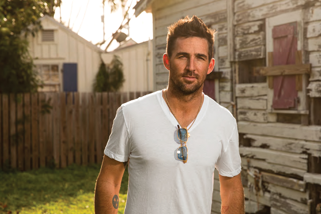 Jake Owen wear a white v-neck T-shirt and a neutral expression. His sunglasses are folded onto his thin necklace.