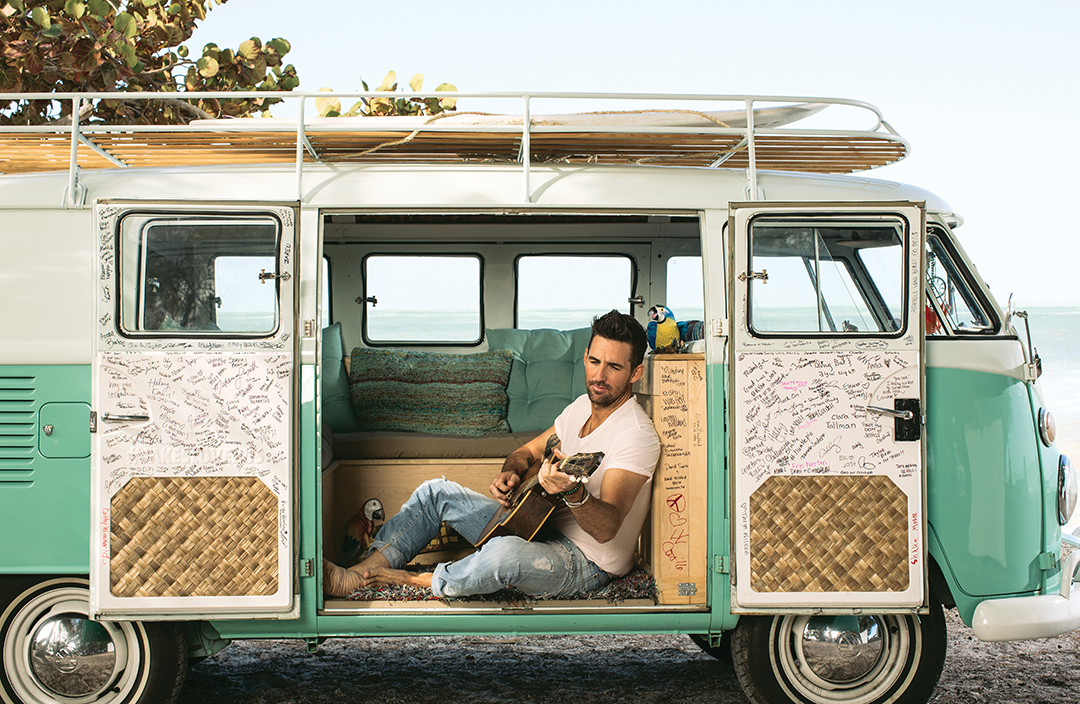 In 2017, Owen drove what he called the “love bus.” In this photo, Owen sits in a vintage van parked at a beach and plays guitar.