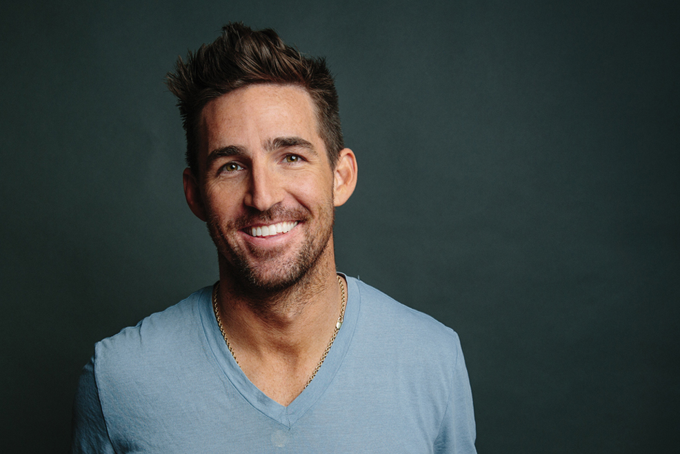 Jake Owen smiles in a portrait. He has light stubble and wears a blue v-neck T-shirt.