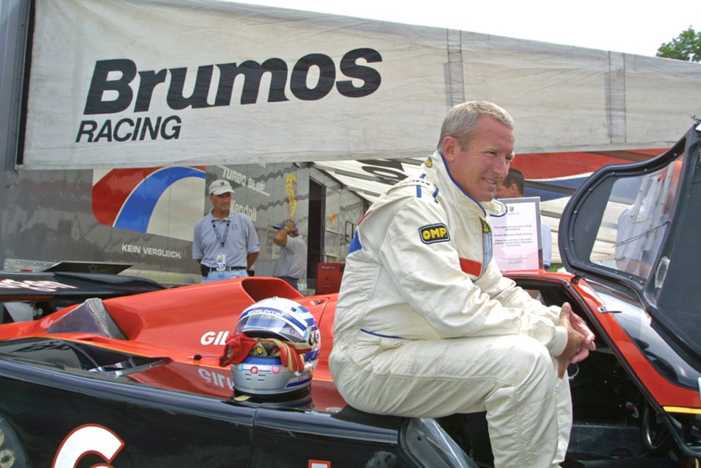 An older Haywood wears a race uniform and sits on the car. In the background, an awning reads Brumos racing. 