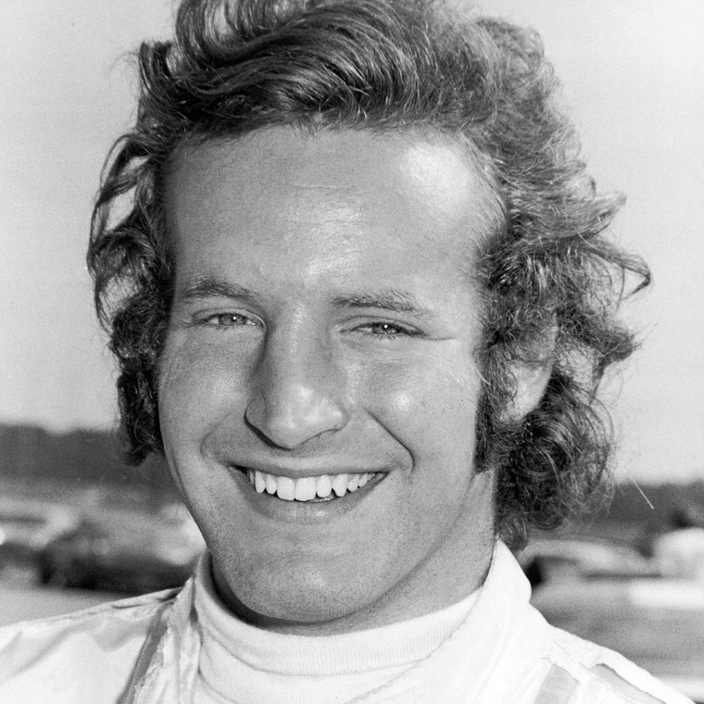 A black and white photo of young, smiling Haywood. His hair is a bit wavy and flows from his hair past his ears and a bit down his neck.