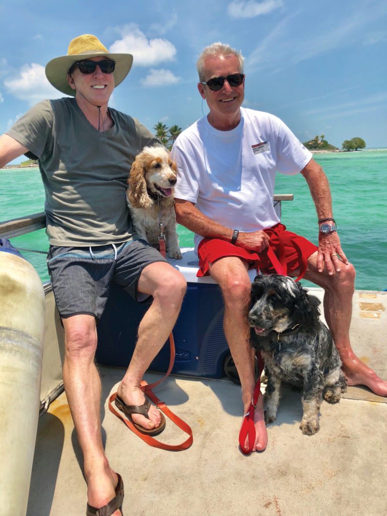 Haywood and his husband and their two dogs smile on a boat. Water is visible in the background.