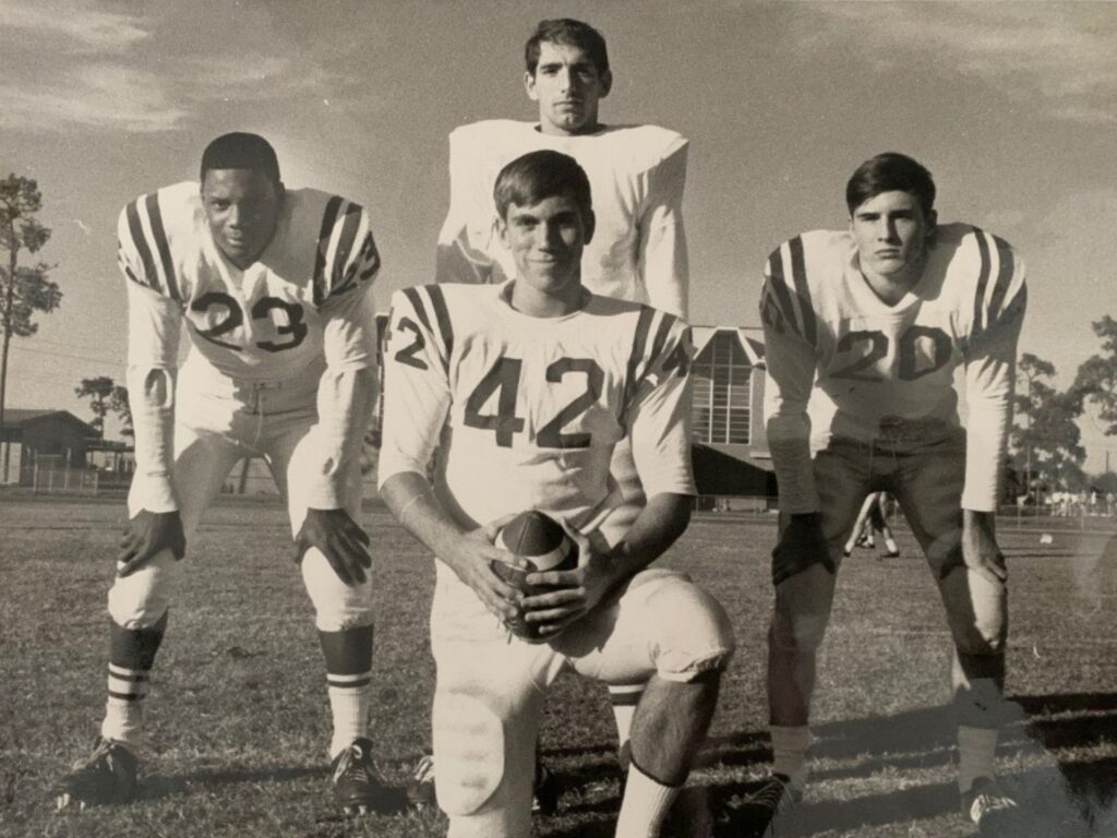 Leonard George, who is Black, crouches with his hands on his knees alongside three white teammates. They all are in football uniforms. George is number 23. 