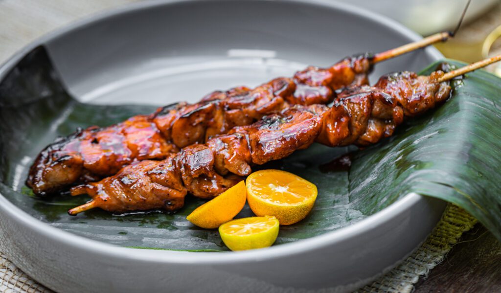 Two kebabs in a gray dish with a citrus garnish and resting on a banana leaf. 