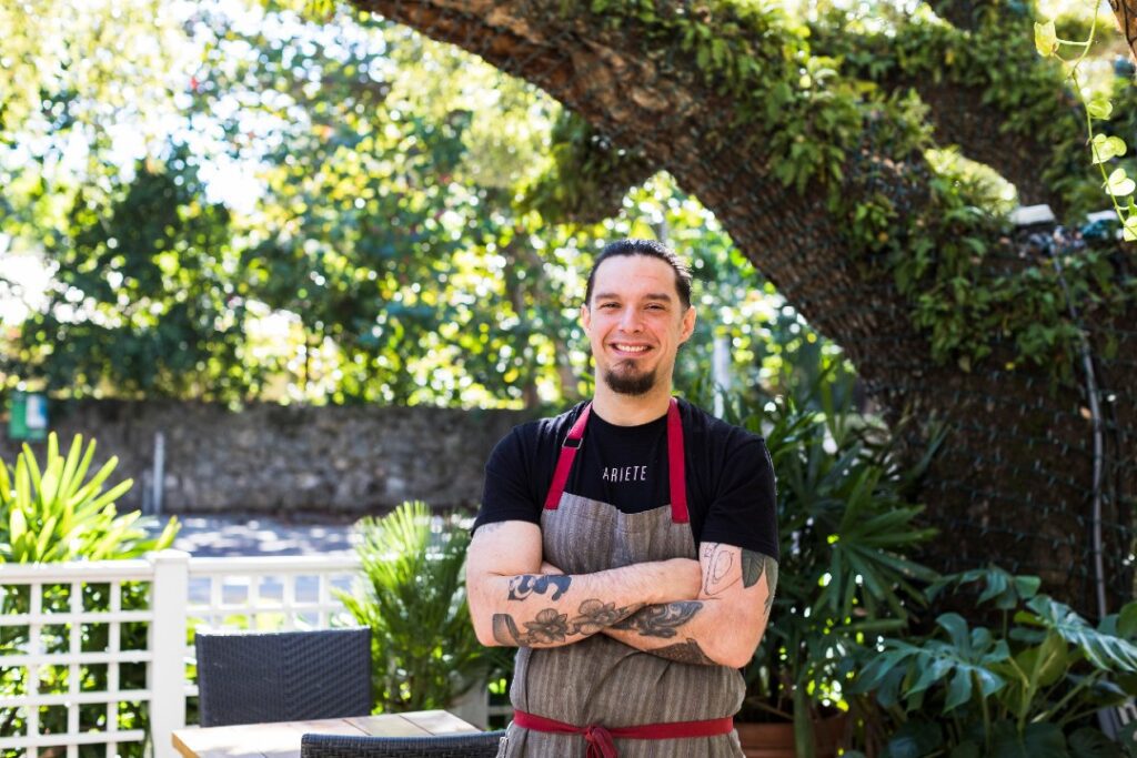 Michael Beltran smiles, arms crossed under a tree. He had tattoos on his arms and is wearing an Ariete T-shirt.